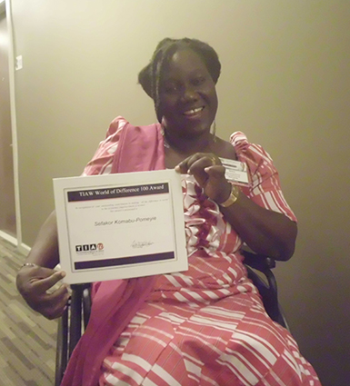 A young woman from Ghana in a wheelchair holding an award that reads TIAW World of difference 100 award.