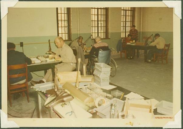 Color photo of a sheltered workshop. 4 people are working on projects, 2 of them are in wheelchairs.